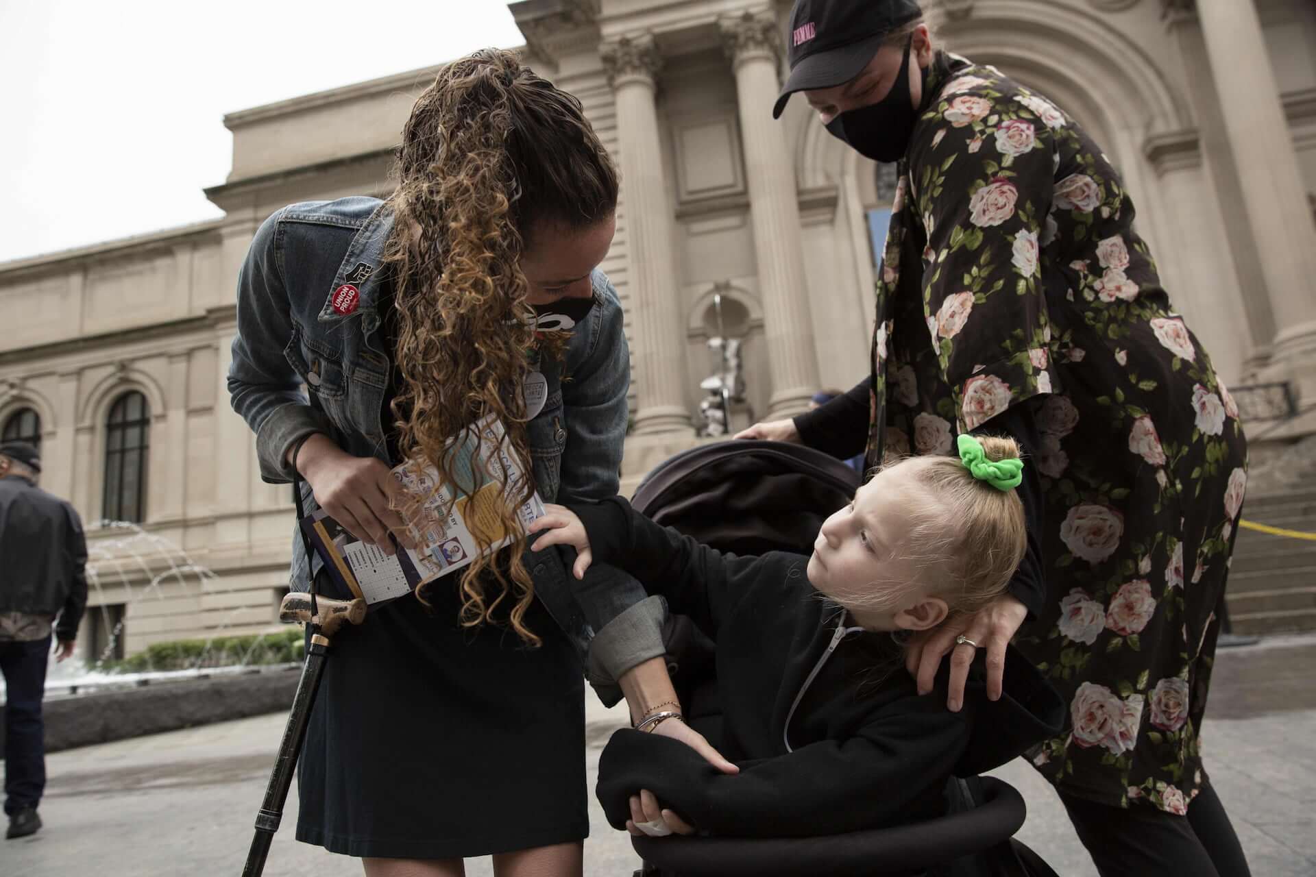 Rebecca bends down to talk to a disabled girl in a stroller and her mother, in front of the Metropolitan Art Museum.