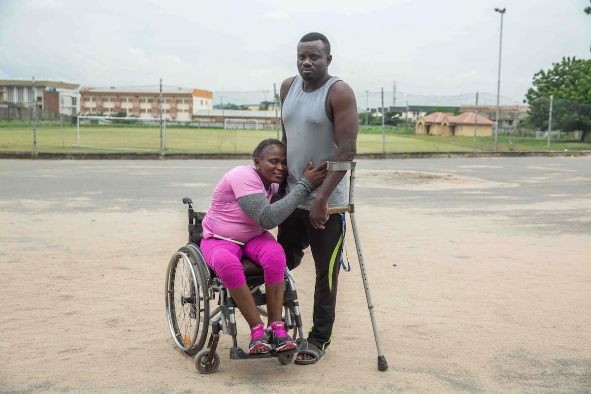 Olajumoke, in her wheelchair, hugs her fiancé Suraju around his waist as they pose for a photo. Suraju has an amputated leg and is using crutches.