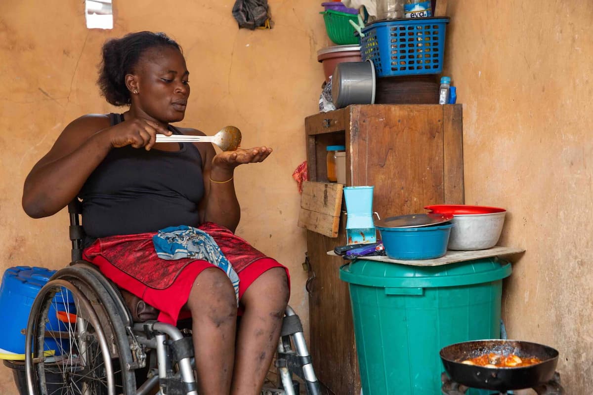Olajumoke at home in her kitchen cooking. She tastes the food she cooks on a single burner, several pans and bowls sit on top of a covered trash can.