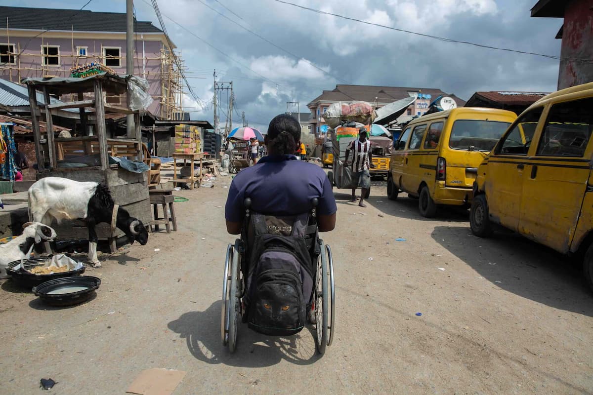 Olajumoke sits in her wheelchair in the middle of a poorly maintained street in Lagos with her back to the camera. The street is crowded with vendors, vehicles, and goats.