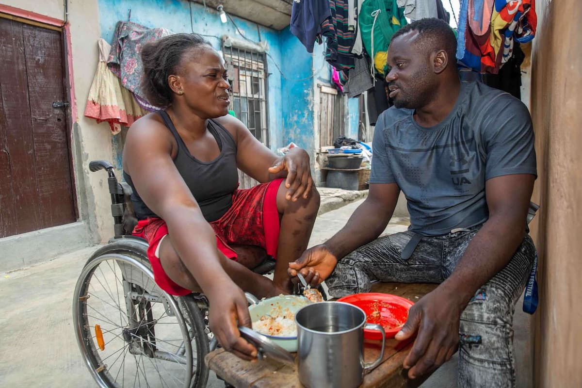 Olajumoke sits in her wheelchair outside her house eating a meal with her fiancé Suraju, in the low-income Amukoko neighborhood.