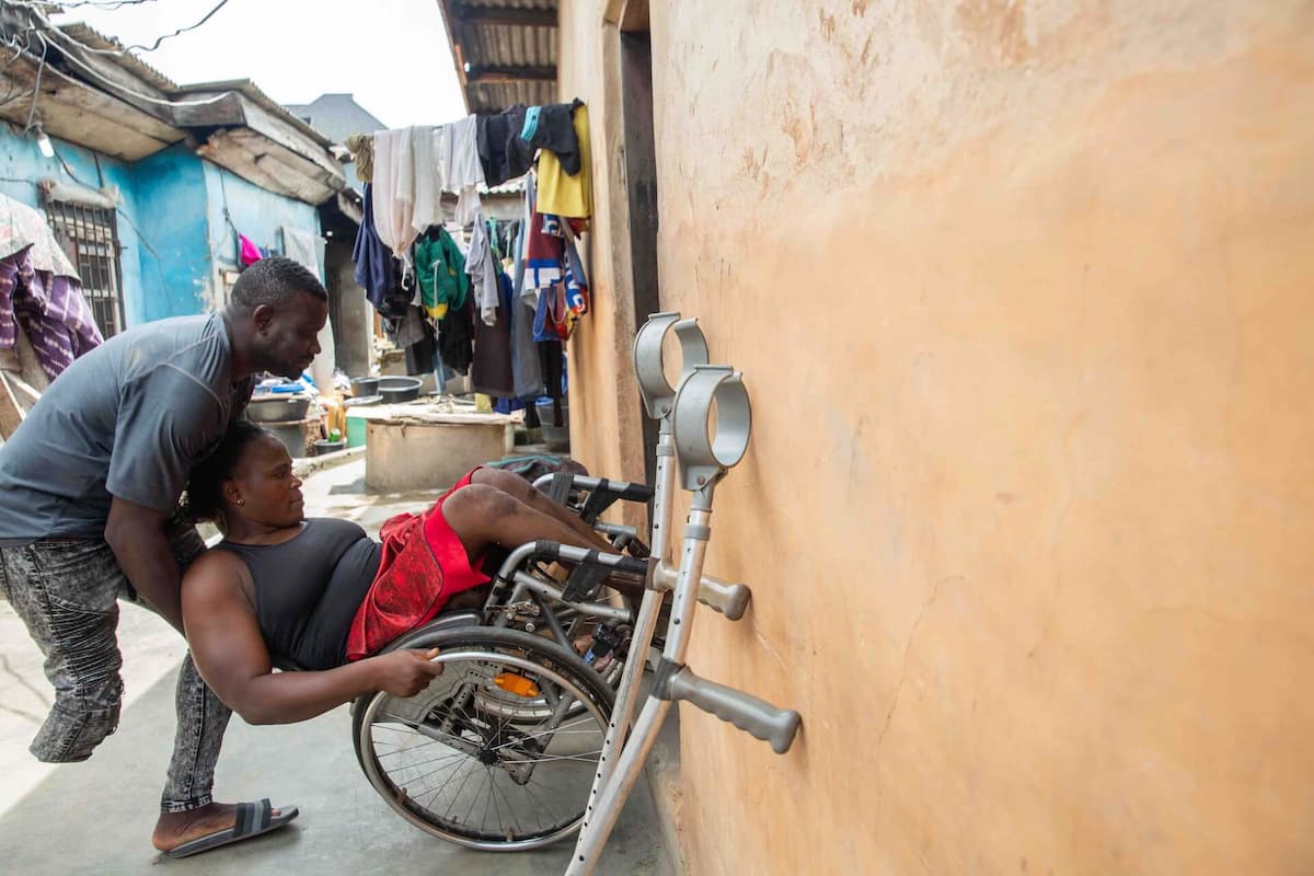 Suraju balances on one leg as he lifts Olajumoke’s wheelchair up a large step into her house. His crutches lean nearby against the building.