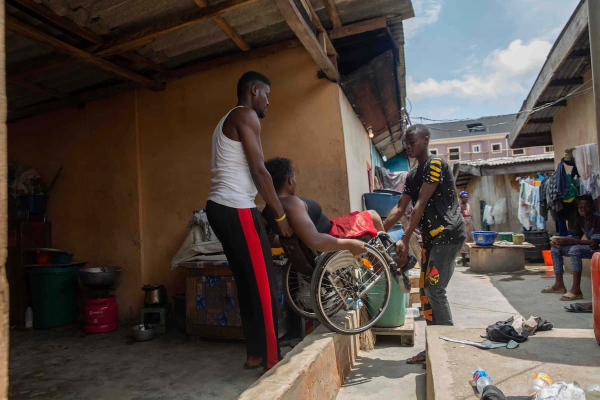 Two men lift Olajumoke’s wheelchair over a high step in her house to get to the alleyway.