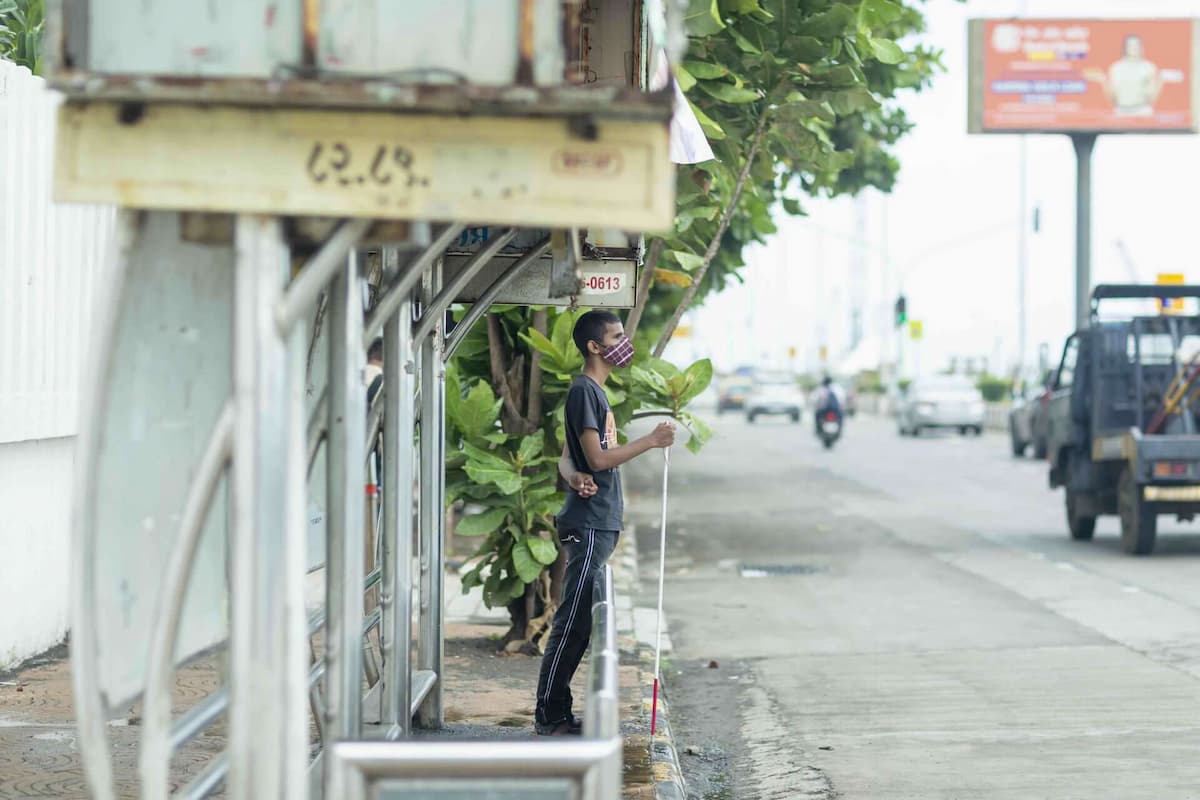 A visually impaired young man holding a white cane leans against a bus stop barrier at the edge of the pavement.