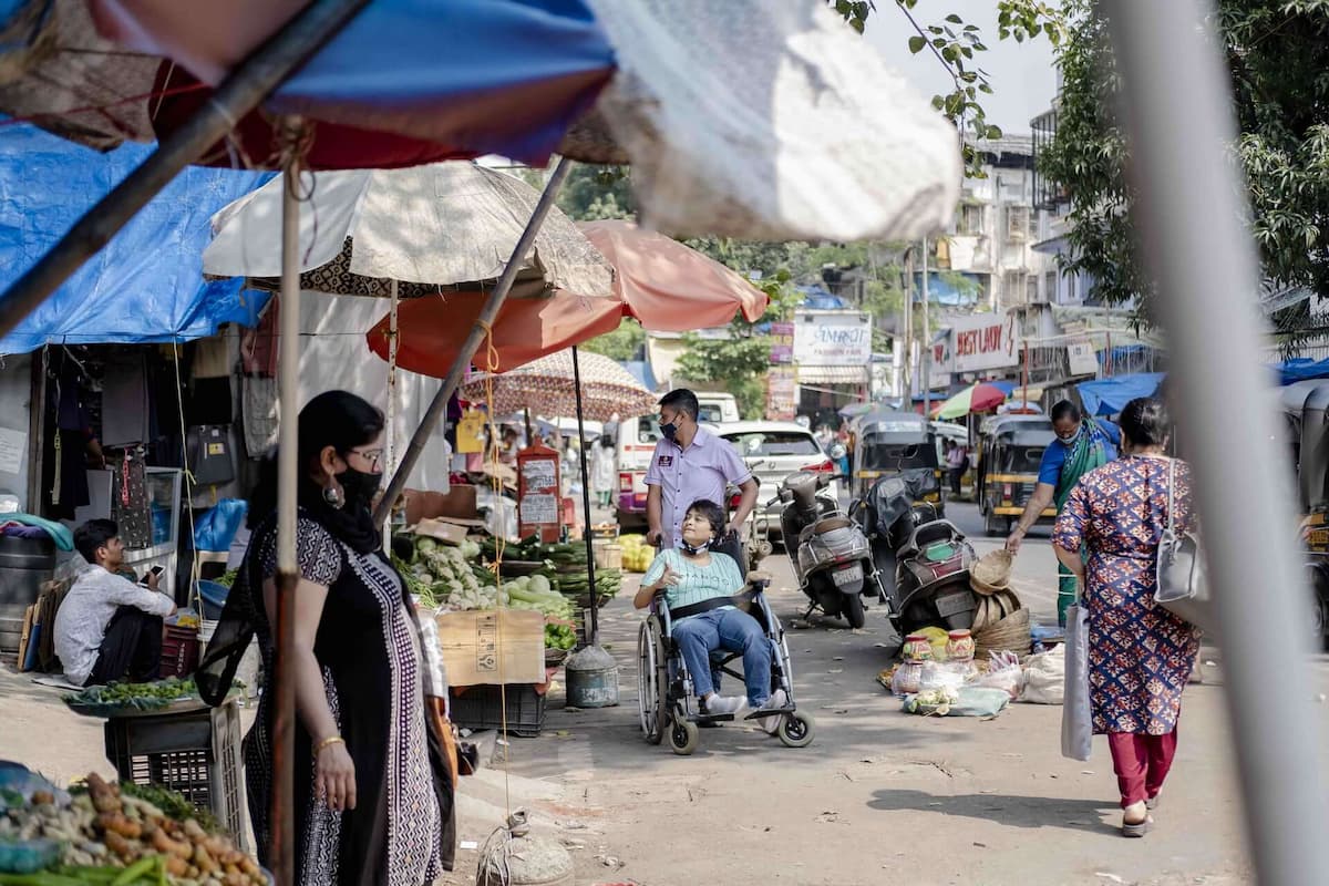 A woman in a wheelchair and her caregiver on a city street crowded with market vendors, people shopping, as well as motorcycles, cars, and vans.