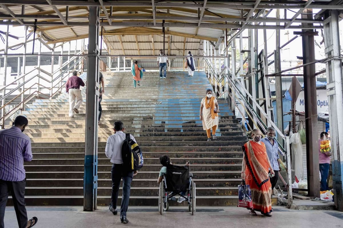 A person in a wheelchair sits at the bottom of a long flight of stairs while people pass her by.