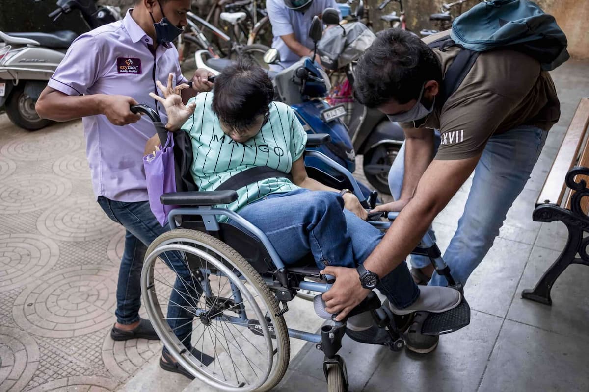 Two men lift a woman with cerebral palsy in a wheelchair up a short flight of stairs.