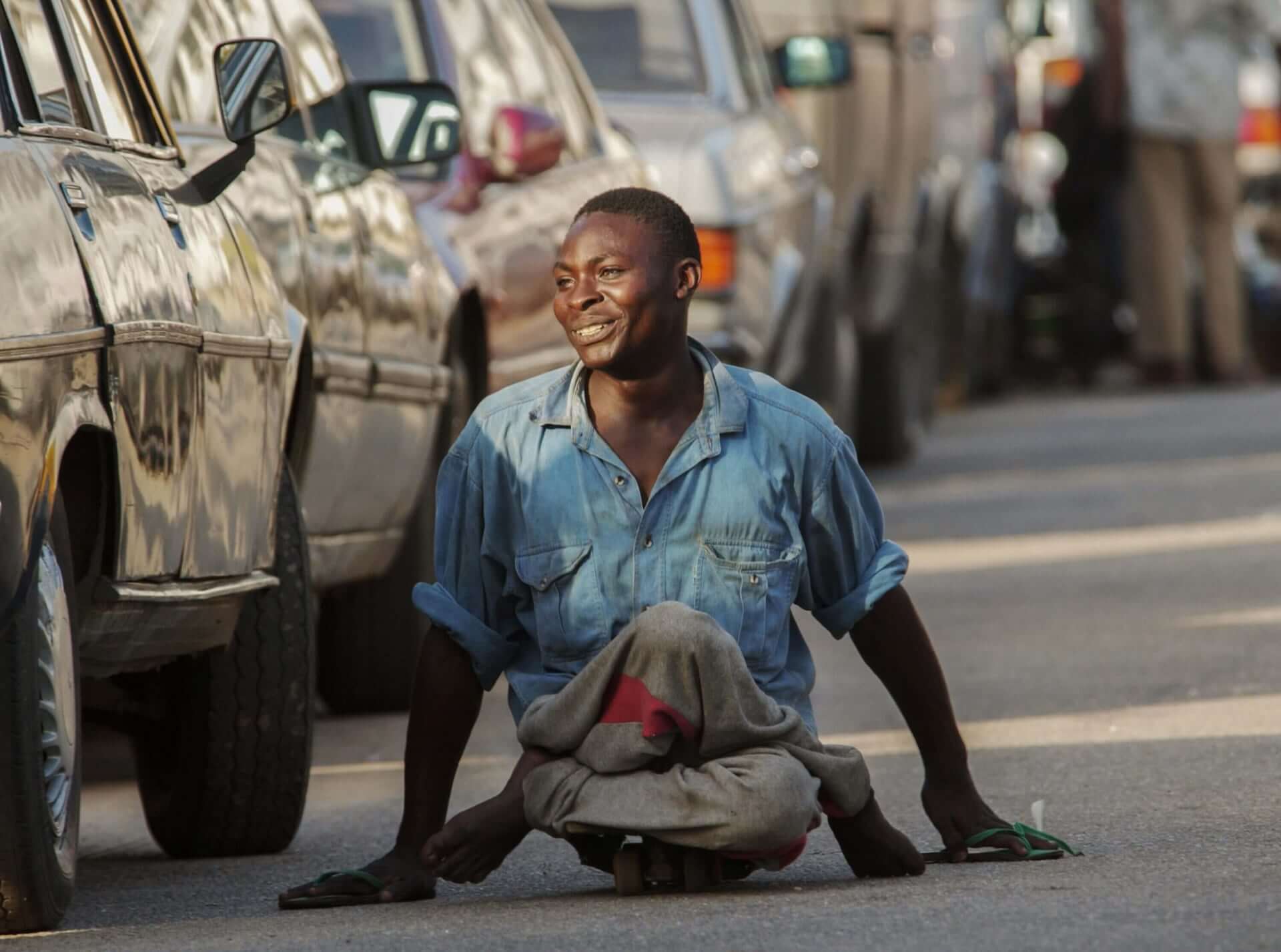 A man crippled by polio rides on a skateboard alongside cars.