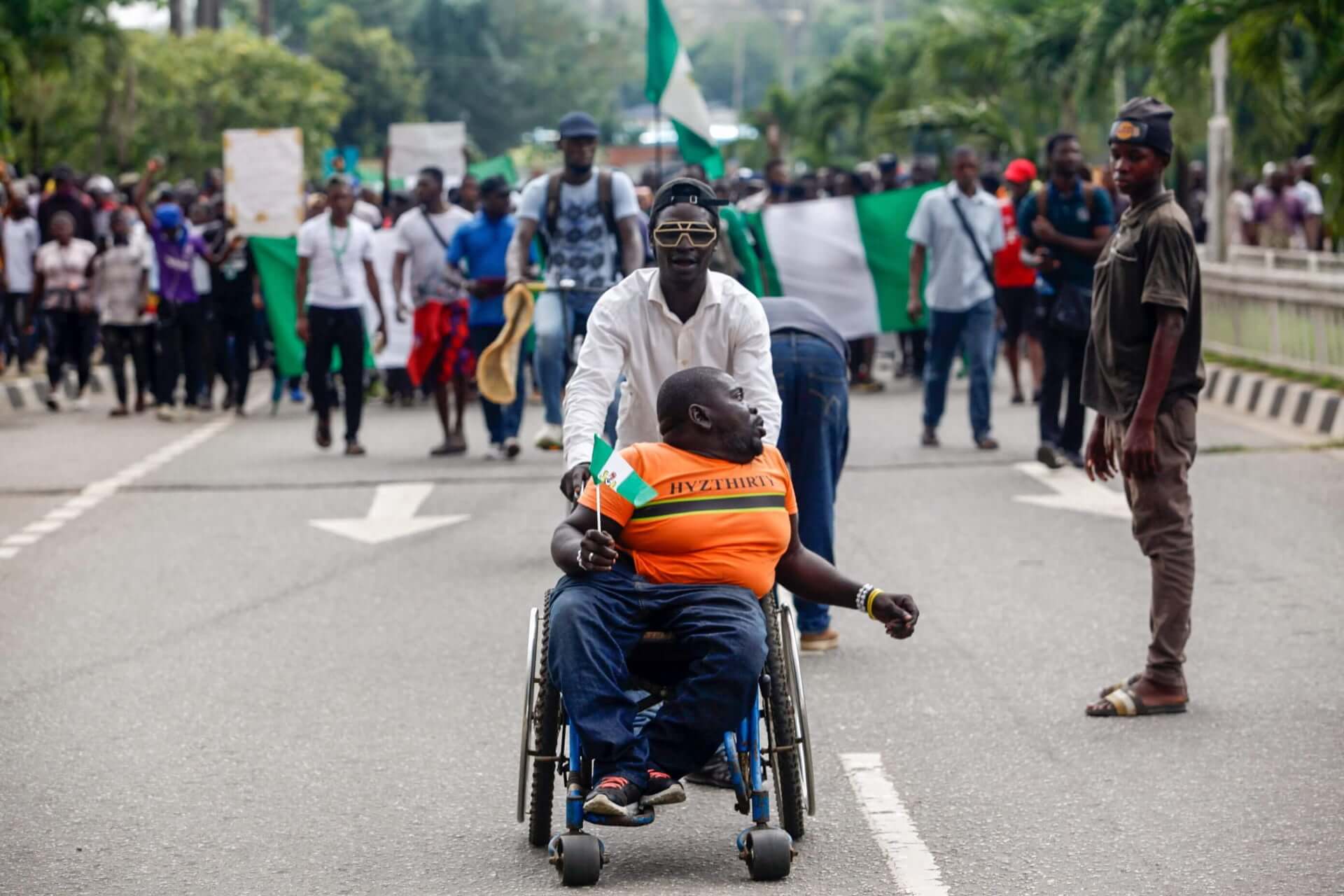 Nigerian demonstrators march behind a man in a wheelchair being pushed by his caregiver.