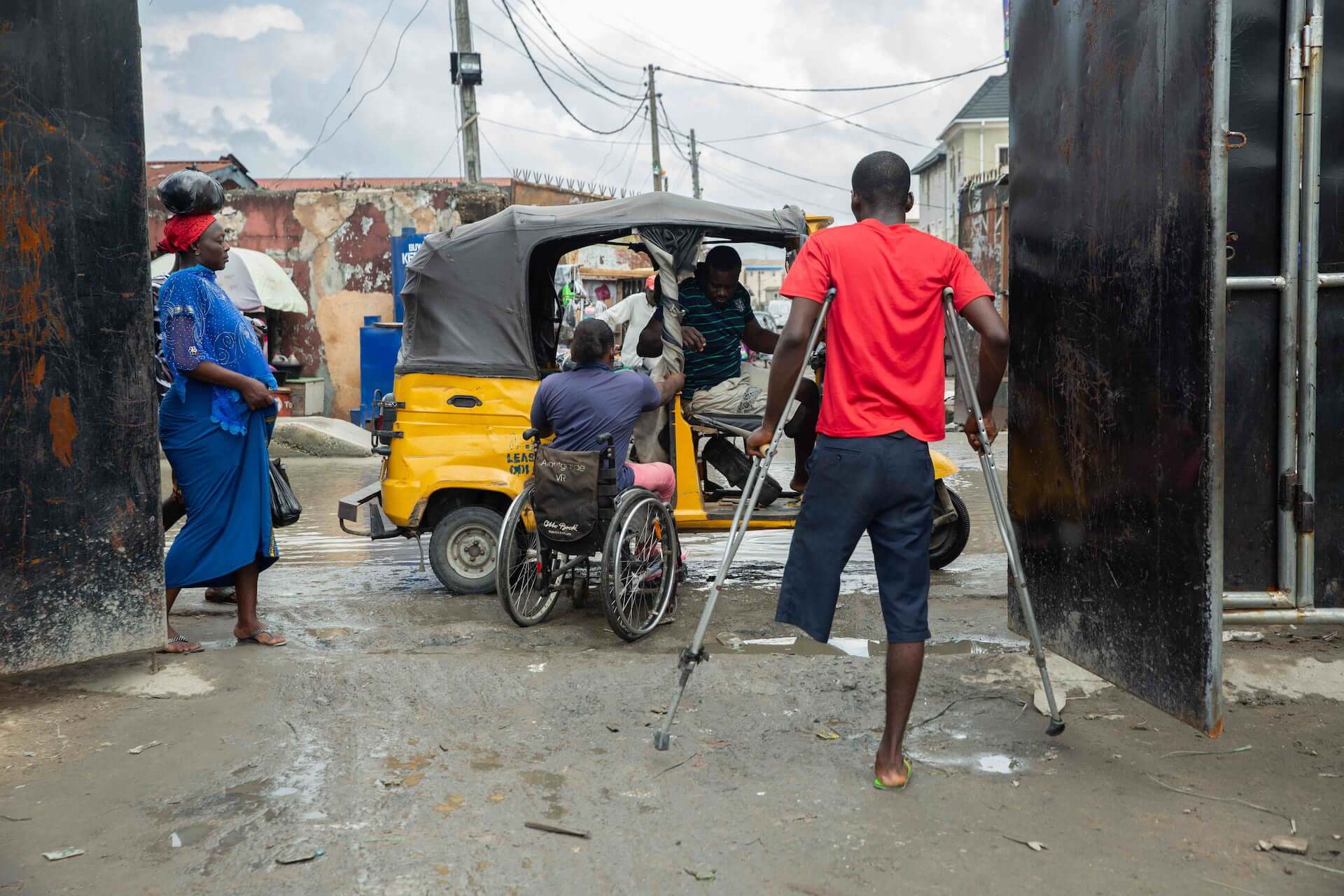 Olajumoke boards an inaccessible keke napep – a yellow and black tricycle. A man with an amputated leg using crutches and a woman watch.