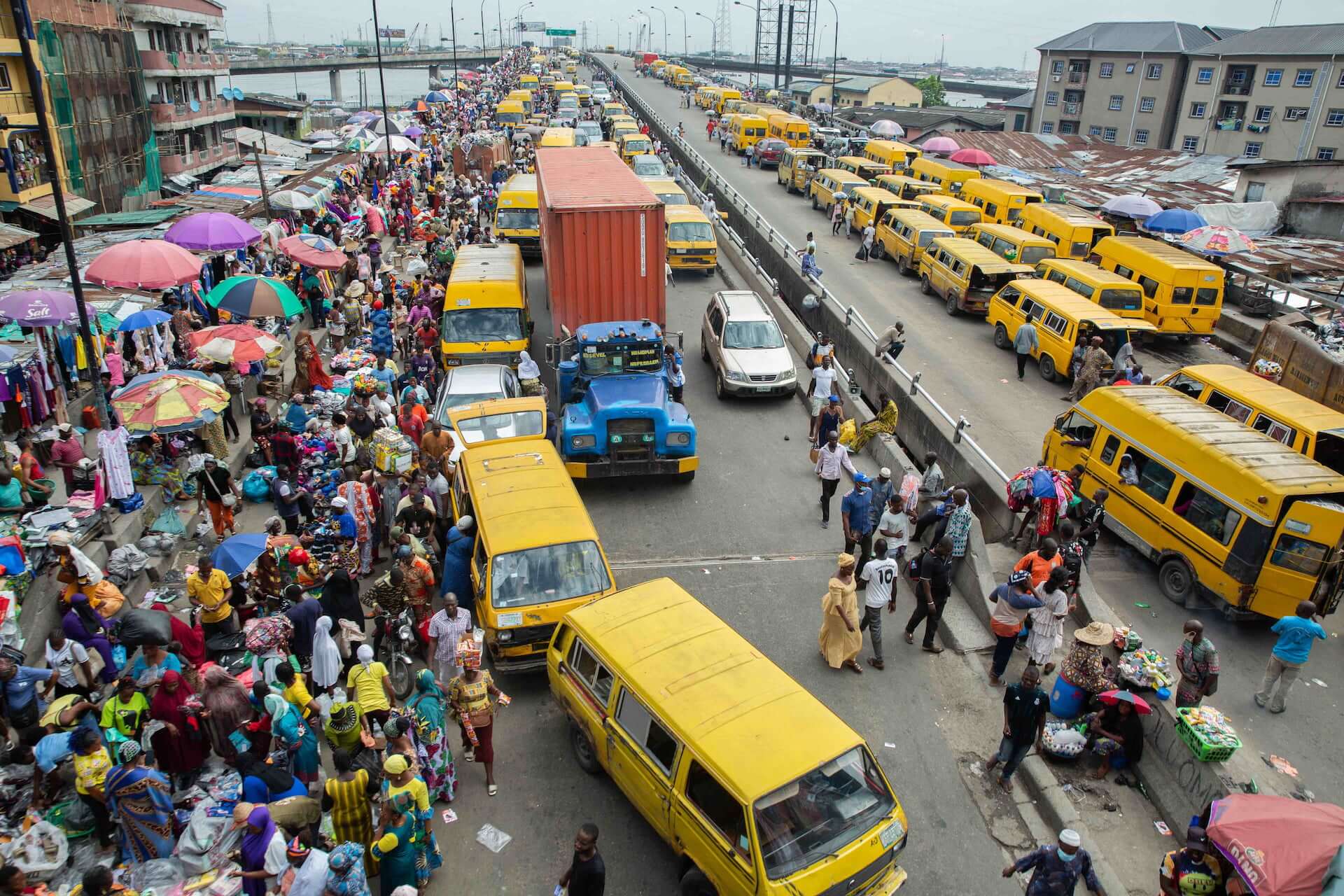 A busy, congested street in Lagos filled with yellow buses and a truck, as well as pedestrians and vendors.