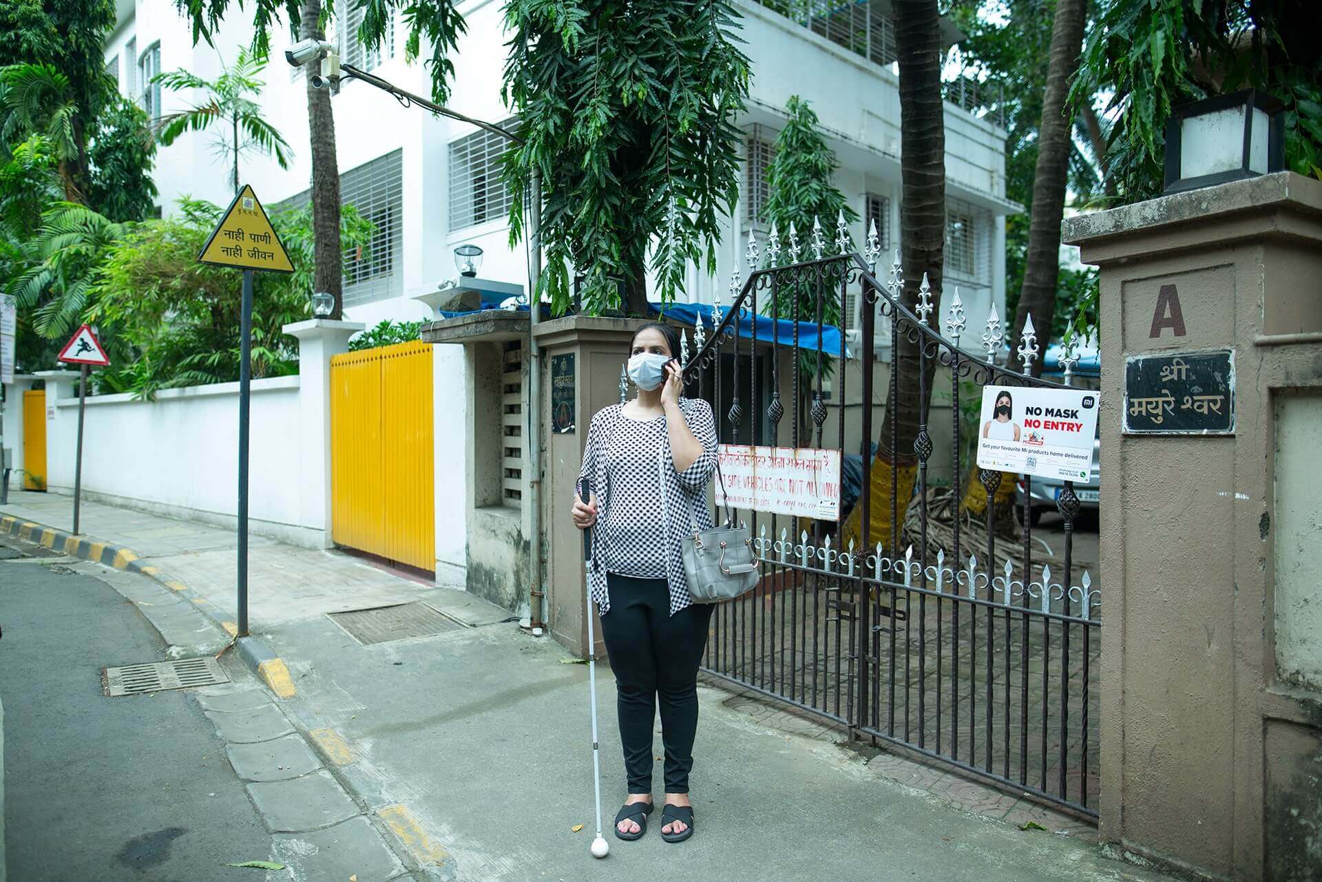 Nidhi Goyal stands outside a gated building in Mumbai holding her white cane and talking on her phone.