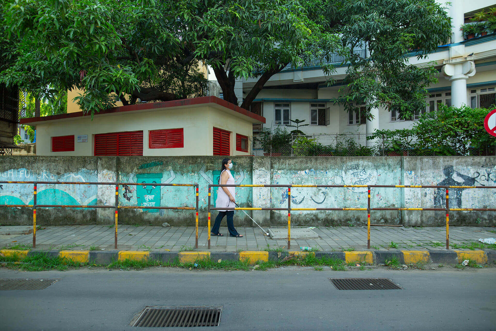 Nidhi walks along a pavement using her white cane. The pavement is separated from local properties by a concrete wall and from the street by a metal fence.