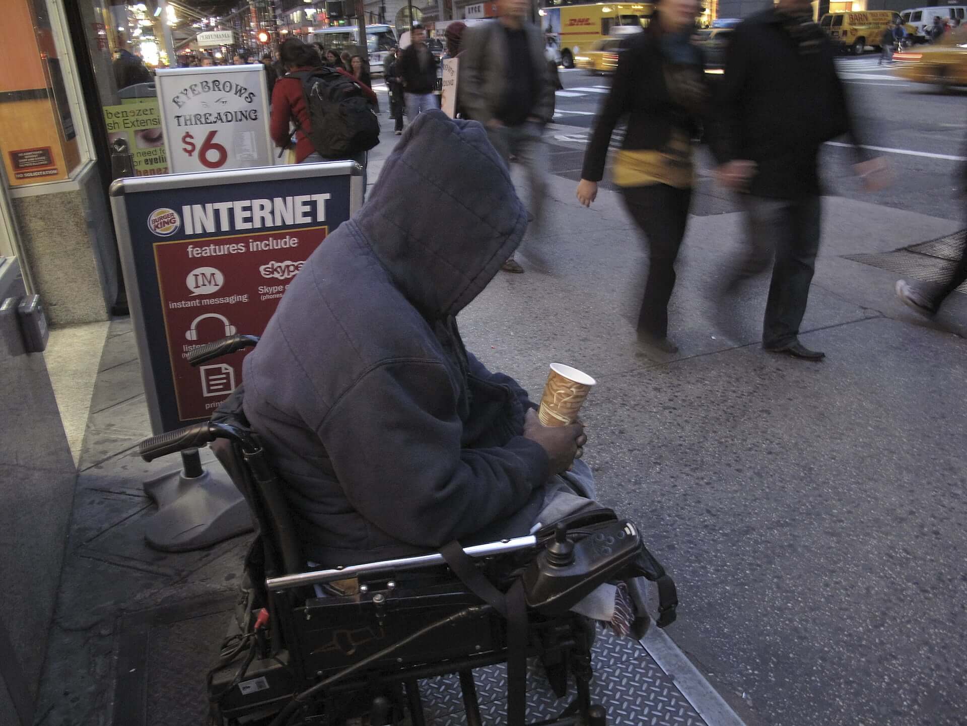 A homeless man with a hoodie covering his head, sits in an electric wheelchair panhandling on a busy New York City street. He holds out an empty coffee cup as people quickly pass by.