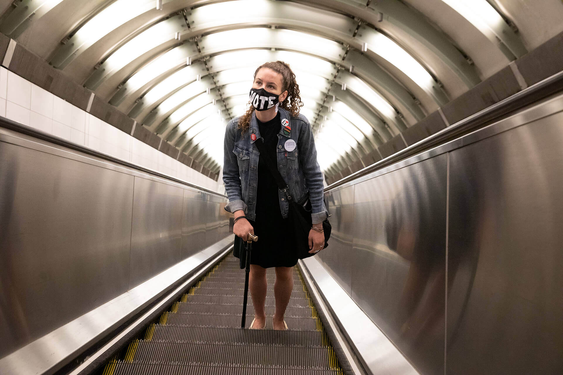 Rebecca rides up a subway station's escalator, wearing her “Vote” mask.