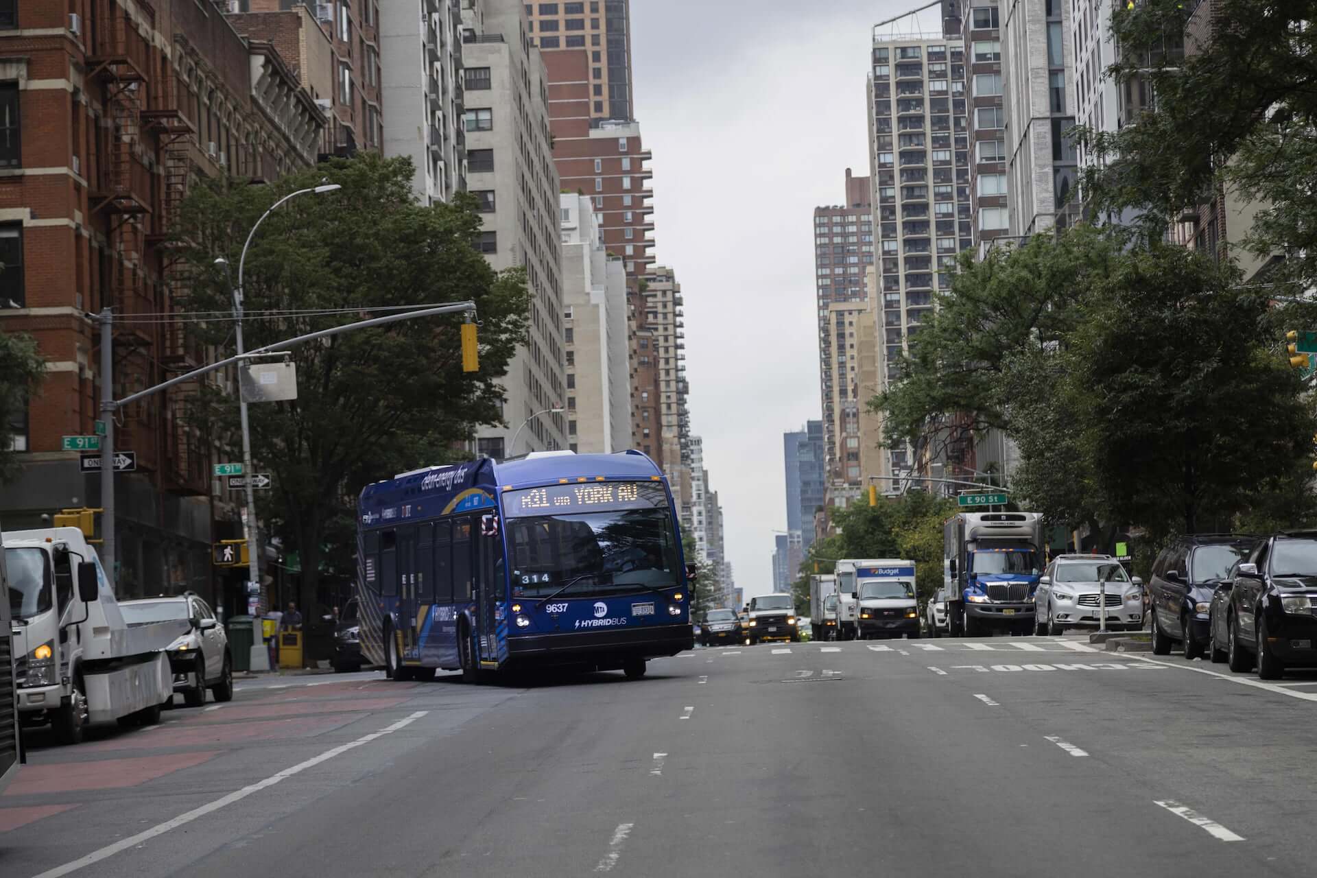 A city bus turns off of East 91st Street to go uptown via 1st Avenue. The street is lined with skyscrapers and green trees.