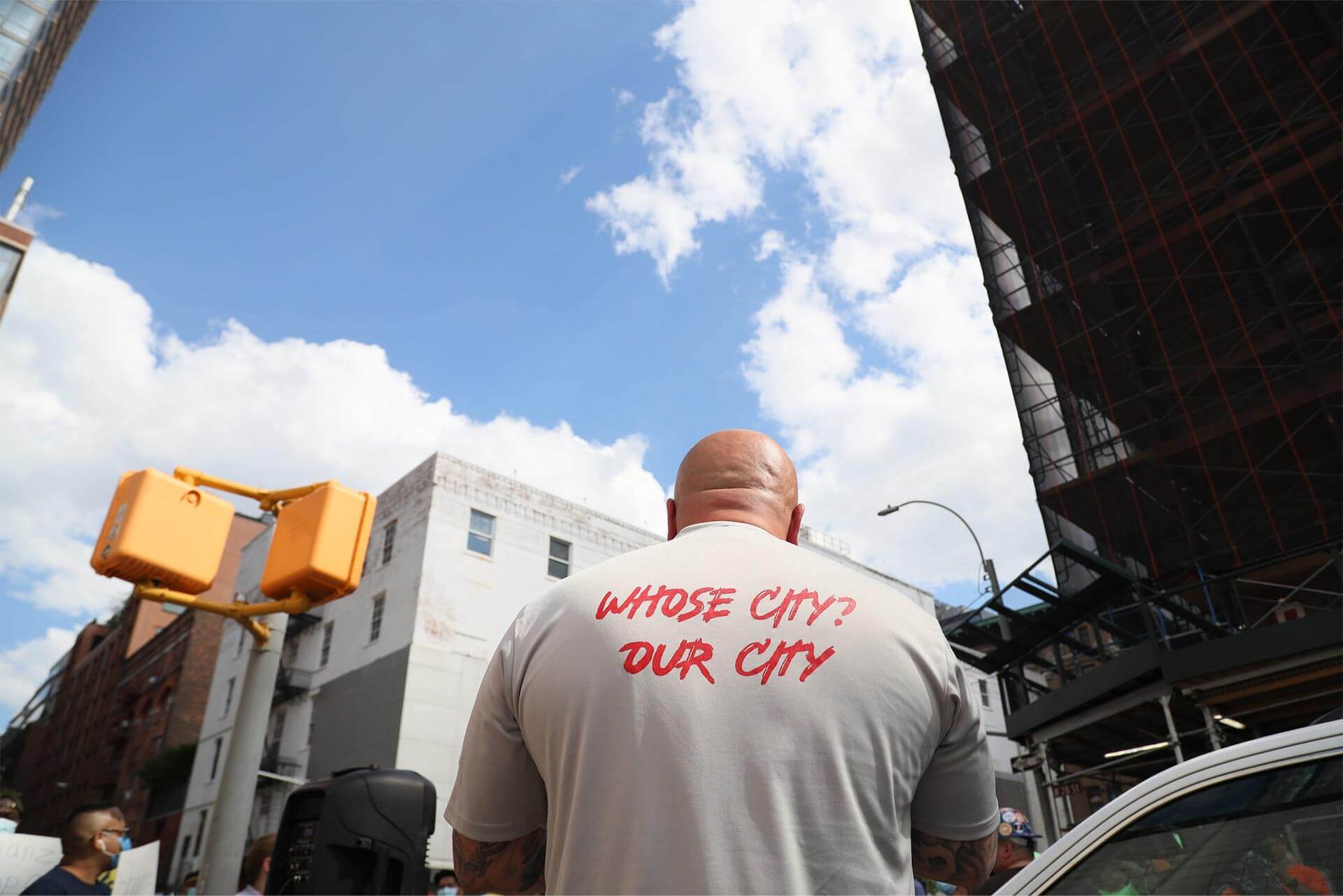 A man wearing a t-shirt with his back to the camera. His shirt reads: Whose city? Our city.